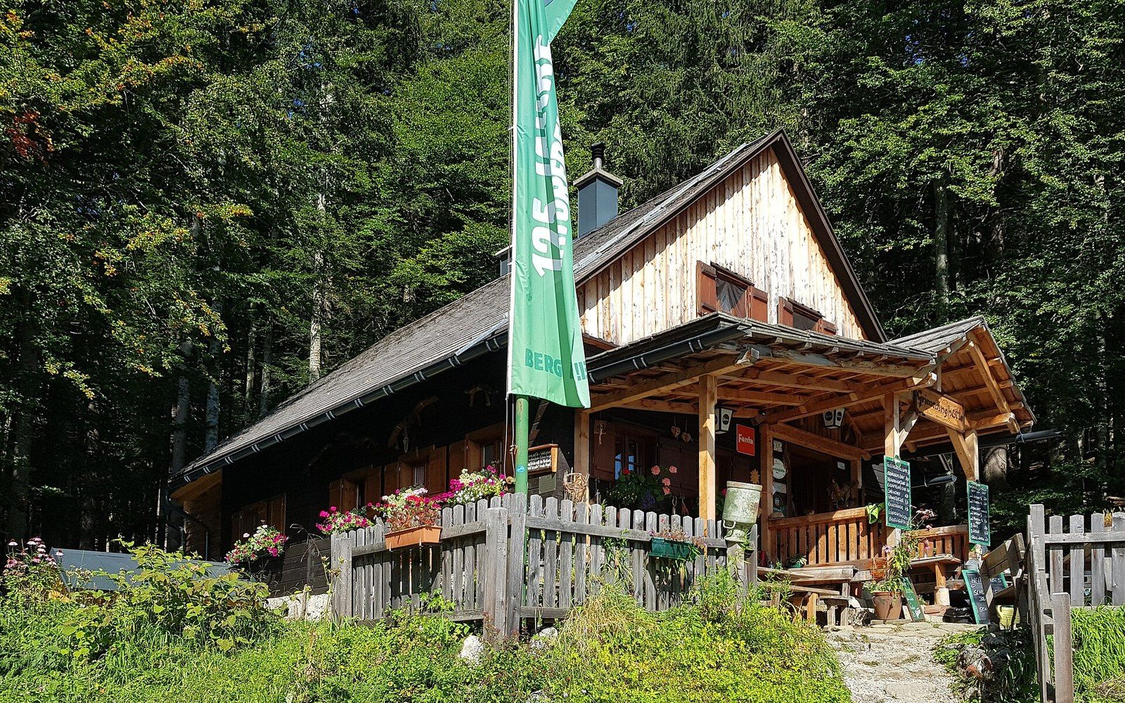 A wooden mountain hut with a flower-lined porch and green flag, surrounded by forest