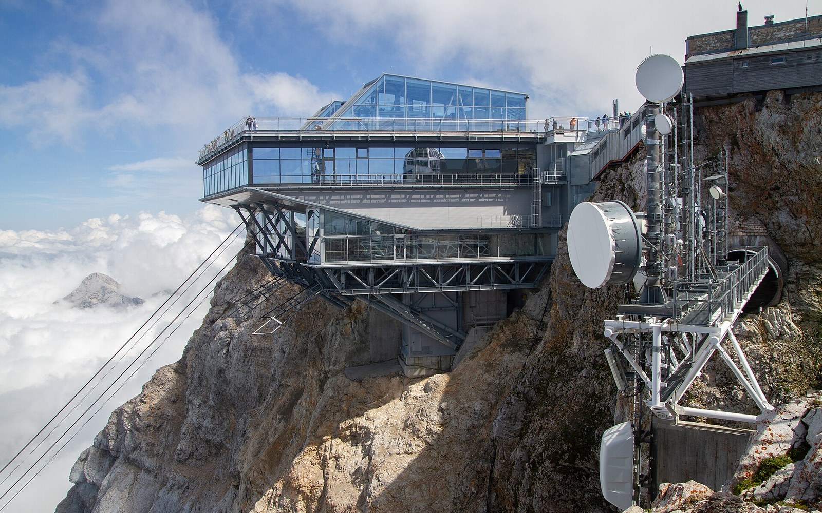A cantilevered glass summit station perches above clouds on a rocky mountain peak